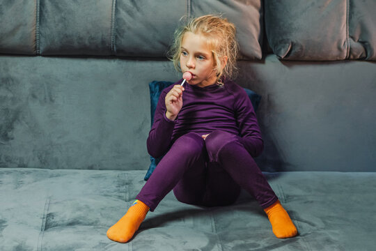 A blonde-haired girl in purple thermal underwear sits brooding on the gray couch in the living room. In her hand is a round lollipop on a stick.