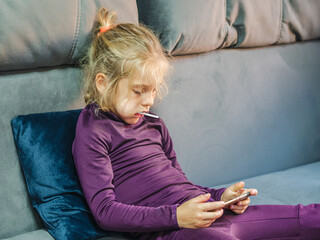 A blond-haired girl with a lollipop in her mouth, watching cartoons on a smartphone on a gray...