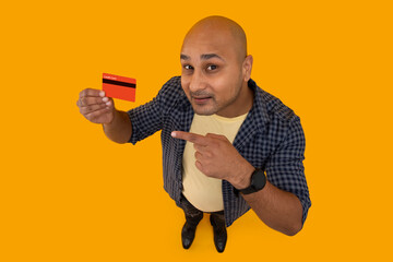 Portrait of a bald man pointing towards the credit card in hand against plain background.