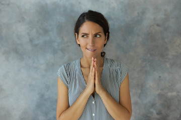 Horizontal indoor shot of suspicious woman frowning with doubt, joining palms together, biting lips, trying to figure out awkward situation, standing against grey and blue studio wall