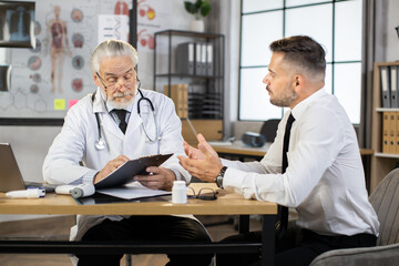 Fototapeta na wymiar Senior bearded doctor in eyewear and lab coat writing on clipboard while listening to male patient in formal clothes. Caucasian businessman visiting competent therapist for health treatment.