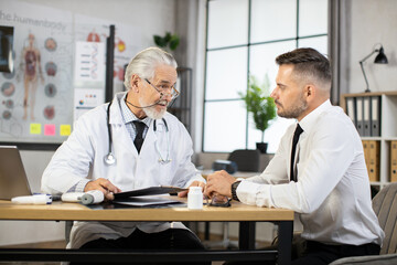 Fototapeta na wymiar Caucasian businessman in formal clothes and senior male doctor in lab coat sitting at medical cabinet and talking. Patient receiving professional consultation and check up a modem clinic.