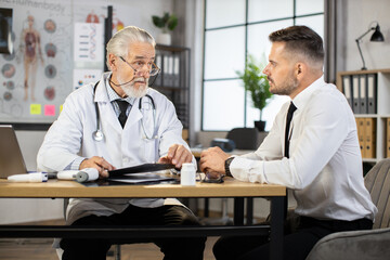 Handsome man in business clothes visiting family doctor for medical consultation. Senior male doctor sitting at desk with patient and talking during appointment. Health care concept.