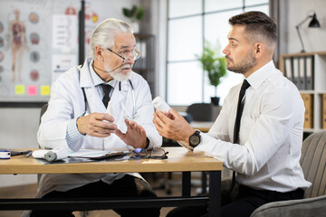Obraz premium Professional male doctor sitting at table with caucasian businessman and giving advices how to take pills. Concept of medication and prescription.