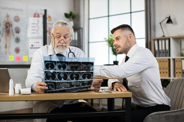 Male patient in business outfit and male doctor in lab coat sitting together at desk and looking at x ray scan. Therapist showing results of tomography to caucasian man.