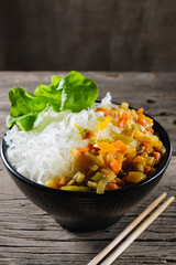 A plate of rice noodles stands on an old wooden table top. The side dish consists of fried onions, carrots, paprika. Vertical