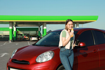 Beautiful young woman with coffee eating hot dog near car at gas station