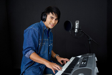 Young asian man with headphones playing an electric keyboard in front of black soundproofing walls....