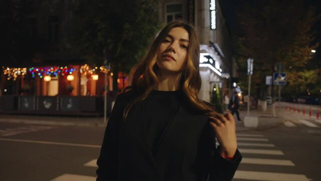 A sensual young girl stands near a pedestrian crossing against the backdrop of colorful garlands and neon lights of the night city. Slow motion