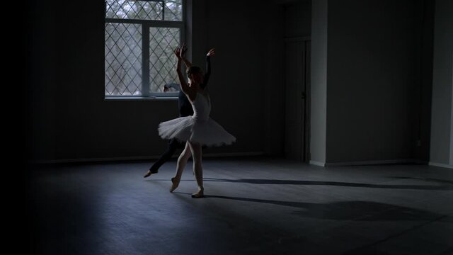 Two flexible ballerinas rehearsing performance in darkness indoors. Wide shot portrait of charming agile Caucasian young women in white tutu and black dress dancing in studio. Art and beauty