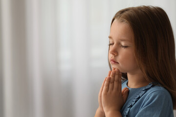 Cute little girl with hands clasped together praying indoors. Space for text