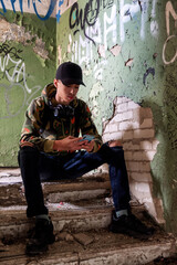 Teenager with black cap sitting on stairs in ruined building and using smarthphone