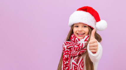 A little girl in a New Year's hat and a warm scarf is very joyful and shows her thumb up. A child in a Christmas costume on an isolated background.