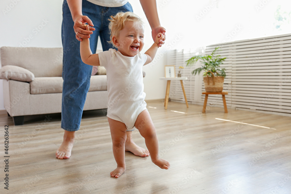 Wall mural first steps of a little girl. a blond toddler learning to walk at home with the help of loving mothe