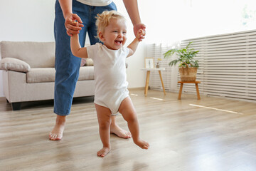First steps of a little girl. A blond toddler learning to walk at home with the help of loving mother. Close up, copy space for text, background. - obrazy, fototapety, plakaty