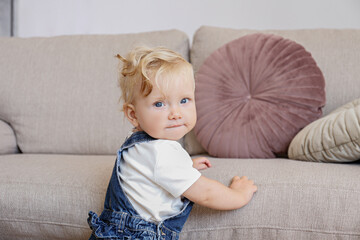 One year old child learning to stand up at living room. Interior background. Adorable blonde little...
