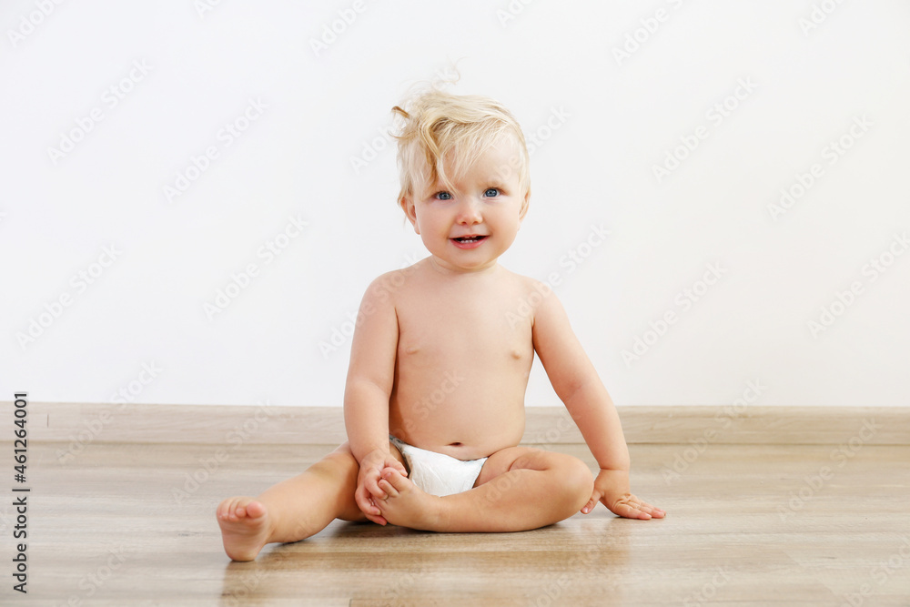 Wall mural Portrait of one year old child sitting on the wooden floor over white wall background. Adorable blonde little girl in diaper. Close up, copy space for text.