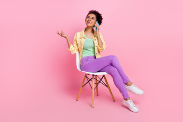 Full length photo of cute young brunette lady sit talk telephone wear yellow shirt jeans shoes isolated on pink background