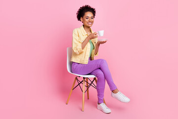 Full size photo of cute millennial brunette lady sit drink tea wear yellow shirt jeans shoes isolated on pink background