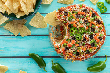 Top down view of a homemade Mexican layered dip served with a bowl of nacho chips. 