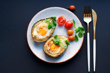 Baked avocado with egg, cheese, corn salad and tomatoes on a white plate on black background. Breakfast with avocado. Gold knife and fork. Top view