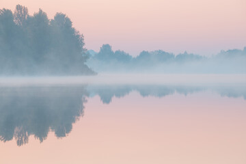 Fototapeta premium Dawn over the foggy lake. Beautiful dreamy view. Pink sky just before the sunrise and fog over water and trees with reflections on the river bank.