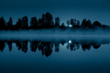 Night mystical scenery. Full moon through the tree branches, rising over the foggy river and its...