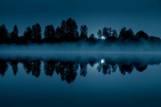 Night mystical scenery. Full moon through the tree branches, rising over the foggy river and its reflection in the still water.