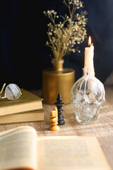 Old books, reading glasses, vintage chess pieces, lit candle and vase with gypsophila flowers. Dark academia concept. Selective focus.