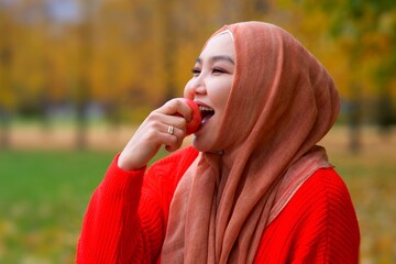 Portrait of happy. beautiful pretty Asian Muslim young woman, Islamic girl is wearing on her head hijab scarf is eating fruit, biting ripe apple. Harvesting. Traditional Muslim Arabian clothes. 