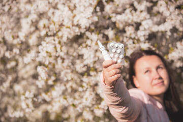 Allergy concept, young woman with pills or drugs and nose or nasal spray against the strong allergy in hand in front of blooming a tree during spring season, healthcare
