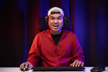 Playing video game. Young asian handsome man sitting on chair in living room. Happiness Streamer Indian man  playing game online wearing headphone in the darkroom.