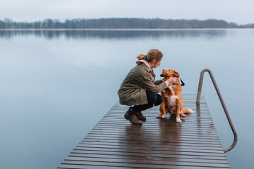 Girl in green jacket sitting on pier with brown Nova Scotia Duck Tolling Retriever and stroking...