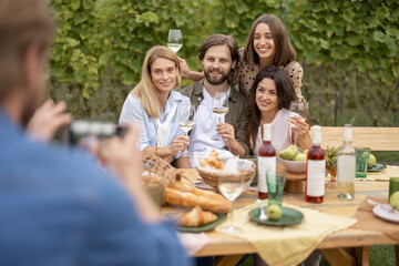Man taking photo on mobile phone of european friends during friendly picnic. Young men and women drinking local wine from glasses. Concept of friendship. Idea of leisure. Green tourism. Winemaking