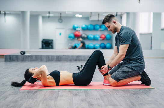 Woman doing abdominal crunches press exercise on the mat with her sports male trainer in gym. Couple exercises on the blurred background of the gym