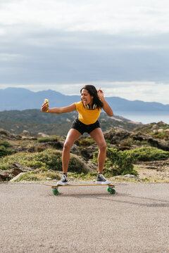 Smiling ethnic woman on longboard and taking selfie