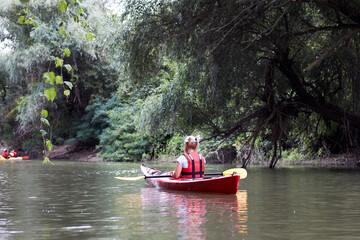 Rear view of woman rowing on red kayak on a Danube river in rural landscape. Summer kayaking in the calm Danube river near the shore with green trees.