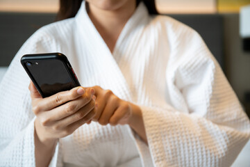 Unrecognizable Asian young woman in bathrobe or shower suit using a smartphone in bedroom.