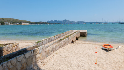 A beautiful beach in the bay of Pollenca on the Mediterranean island of Mallorca. Many boats are tied to buoys in front of the beach. Mountains can be seen in the background.