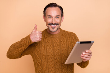 Photo of optimistic brunet man thumb up hold tablet wear knitwear sweater isolated on beige color...
