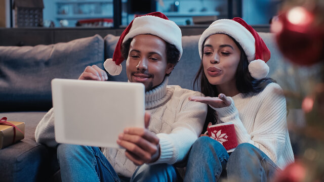 happy african american man near woman sending air kiss and looking at digital tablet during video call