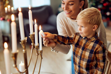 Happy mother assists her small son to light up candles in menorah during Hanukkah celebration.
