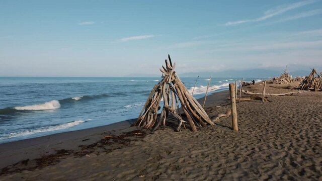 Slow cinematic aerial drone footage of flying away from a wooden driftwood tipi at a sandy beach at the seaside near Alberese in the iconic Maremma nature park in Tuscany, Italy, with blue waves.