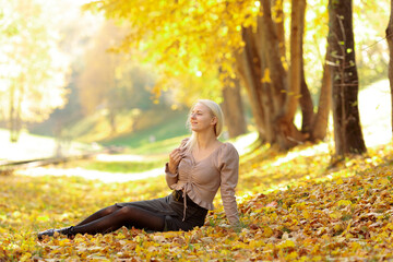 blonde girl in street clothes sitting in a heap of autumn leaves