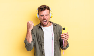 young handsome man shouting aggressively with an angry expression headphones and smartphone concept