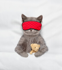Cozy kitten wearing sleeping mask sleeps with toy bear under white blanket on a bed at home. Top...