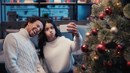Fototapeta premium happy african american man taking selfie with woman puffing cheeks near christmas tree