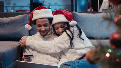 happy african american couple in santa hats hugging while online shopping near blurred christmas...