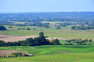 A view of a vast field, meadow, or pastureland surrounded with some trees, shrubs and other flora with some foest or moor in the distance and some tractor ploughing the field seen in Poland