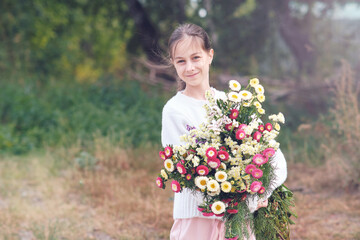 A girl holds in her hands a large beautiful bouquet of autumn flowers in a white sweater in a forest glade